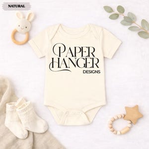 Puede incluir: Body de beb&eacute; color crema con el texto "PAPER HANGER DESIGNS" en negro. La imagen incluye un sonajero de conejito de madera, un par de calcetines blancos para beb&eacute; y un mordedor en forma de estrella de madera. Una ramita de vegetaci&oacute;n est&aacute; en el fondo.