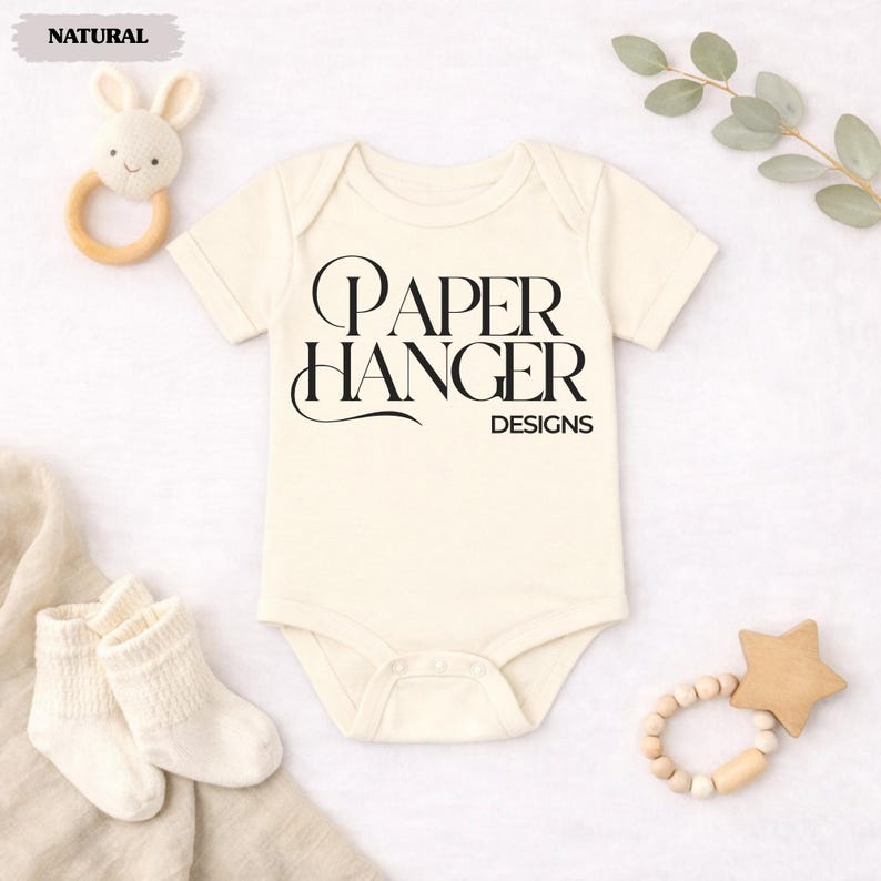 Puede incluir: Body de beb&eacute; color crema con el texto "PAPER HANGER DESIGNS" en negro. La imagen incluye un sonajero de conejito de madera, un par de calcetines blancos para beb&eacute; y un mordedor en forma de estrella de madera. Una ramita de vegetaci&oacute;n est&aacute; en el fondo.