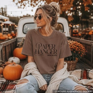 Könnte beinhalten: Ein braunes kurzärmeliges T-Shirt mit dem schwarzen Schriftzug PAPER HANGER MOCKUPS auf der Vorderseite. Das T-Shirt hat einen Rundhalsausschnitt und wird mit einer hellblauen, zerrissenen Jeans und einer cremefarbenen Strickjacke kombiniert.