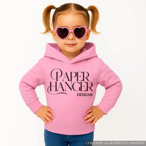 May include: A young child wearing a light pink hoodie with the text "PAPER HANGER DESIGNS" in black. The child is also wearing pink heart-shaped sunglasses and blue jeans. The image is set against a white background.