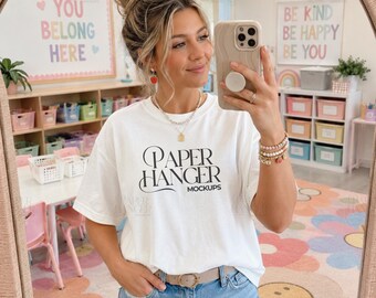Pode incluir: Uma t-shirt branca de manga curta com o texto preto Paper Hanger Mockups impresso na frente. A peça é combinada com calças de ganga azuis presas por um cinto castanho claro com fivela circular e uma pulseira de contas no pulso.