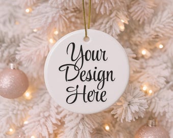 Könnte beinhalten: Ein weißer, runder Keramik-Weihnachtsschmuck mit dem Text "Your Design Here" in schwarzer Schrift. Der Anhänger hängt an einem goldenen Faden vor einem weißen Weihnachtsbaum mit warmen Lichtern und rosa Kugeln.