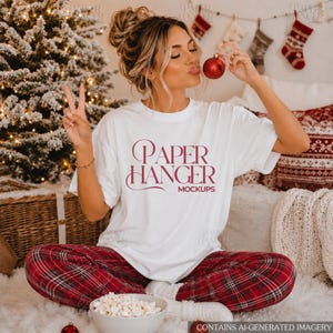 May include: A white short-sleeved t-shirt featuring the maroon text PAPER HANGER MOCKUPS printed on the front. The shirt is worn with red and black plaid pajama pants. The design includes a stylized font with decorative flourishes on the letters.