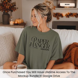 Pode incluir: Uma t-shirt de manga curta verde-musgo com o texto preto "PAPER HANGER MOCKUPS" estampado na frente. A pe&ccedil;a tem gola redonda e um corte descontra&iacute;do. O texto est&aacute; escrito numa fonte serifada estilizada com elementos decorativos.