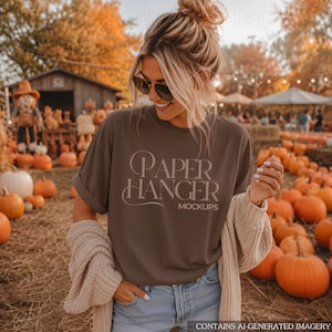 以下が含まれることがあります： 「PAPER HANGER MOCKUPS」という文字が明るいセリフ体でプリントされたダークブラウンの半袖Tシャツ。ベージュのニットカーディガンとライトブルーのデニムジーンズを合わせ、背景にはカボチャ畑が見える。