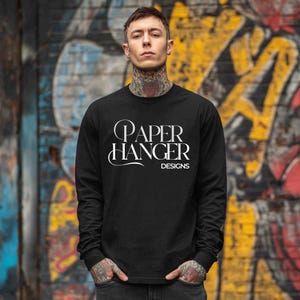 May include: Black long-sleeved shirt featuring the white text "PAPER HANGER DESIGNS". The shirt is worn by a person with tattoos, set against a colourful graffiti-covered wall.
