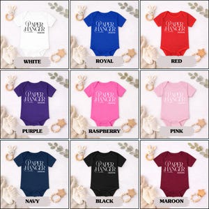 Puede incluir: Una colecci&oacute;n de bodies para beb&eacute;s en varios colores: blanco, azul real, rojo, morado, frambuesa, rosa, azul marino, negro y granate. Cada body presenta el texto "PAPER HANGER DESIGNS".