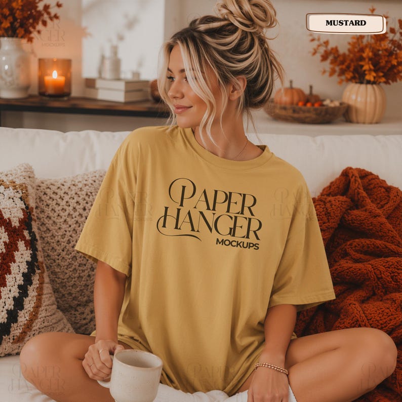 Pode incluir: Uma t-shirt de manga curta amarelo-mostarda com o texto preto "PAPER HANGER MOCKUPS" estampado na frente. A pe&ccedil;a tem gola redonda e um corte descontra&iacute;do, com a palavra "MUSTARD" exibida numa pequena caixa retangular no topo.