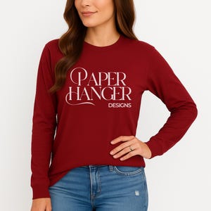 May include: A long-sleeved, burgundy shirt with the text "PAPER HANGER DESIGNS" in white script. The shirt is paired with blue jeans. The model is wearing a ring.