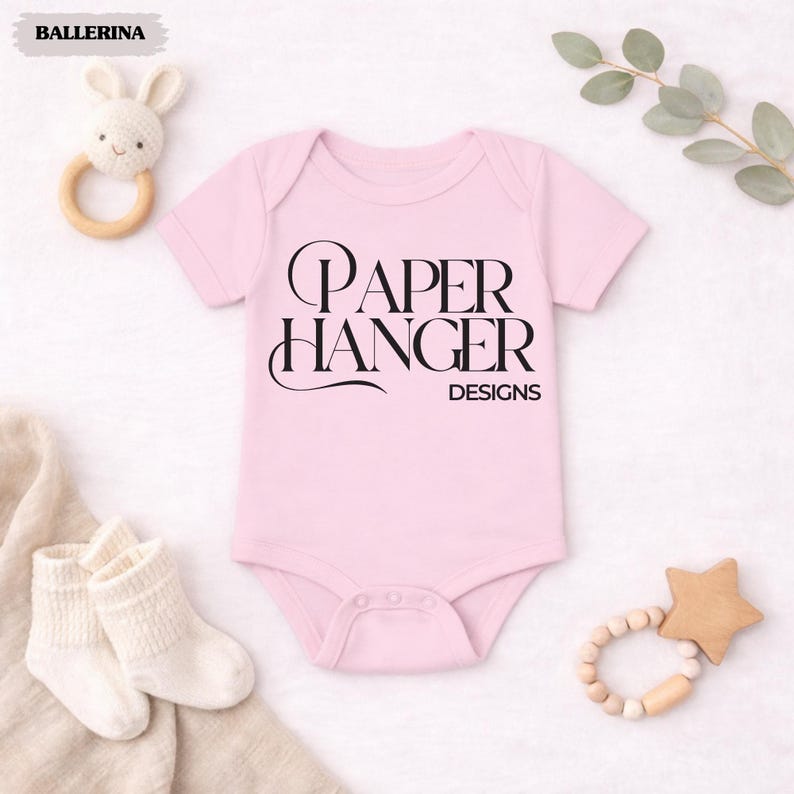 Puede incluir: Un body de beb&eacute; rosa claro con el texto "PAPER HANGER DESIGNS" en escritura negra. La imagen incluye un sonajero de conejo blanco, calcetines de beb&eacute; blancos y un mordedor en forma de estrella de madera. La palabra "BALLERINA" est&aacute; en la esquina superior izquierda.