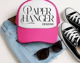 Könnte beinhalten: Eine pinkfarbene und weiße Trucker-Mütze mit dem Text "PAPER HANGER DESIGNS" in Schwarz. Die Mütze wird mit einer schwarzen Sonnenbrille, Jeansshorts und schwarzen Leinenschuhen kombiniert.