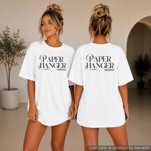 May include: A white short-sleeved t-shirt featuring the black text Paper Hanger Designs printed on the front and back. The shirt has a crew neckline and a loose, relaxed fit, showcasing the brand name in a stylized serif font with flourishes.