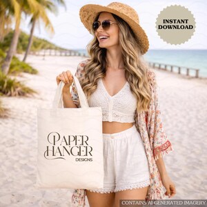 Puede incluir: Una mujer con sombrero de paja y gafas de sol sostiene una bolsa de mano crema con el texto "PAPER HANGER DESIGNS" en negro. Lleva un top de encaje blanco, pantalones cortos y un kimono floral en una playa.