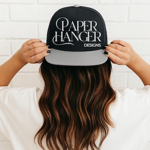 May include: Black and gray baseball cap with the text "PAPER HANGER DESIGNS" in white script. The cap has a flat brim and a button on top. The person is wearing a white shirt.
