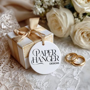 Op de afbeelding: Een witte geschenkdoos met gouden lint en een label met de tekst "PAPER HANGER DESIGNS". Twee gouden trouwringen liggen naast de doos. Witte rozen en kant maken de elegante scène compleet.