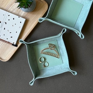 Op de afbeelding: Twee lichtblauwe leren dienbladen met gevouwen randen. Een dienblad is leeg en het andere dienblad bevat een gouden haarspeld en twee gouden ringen.