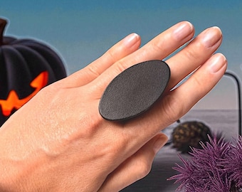 May include: A black oval ring is worn on a hand, with a Halloween theme. The background includes a carved pumpkin, candles, and decorative elements. The ring is the focal point, with a smooth, dark surface.