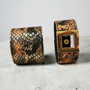 Pode incluir: Duas pulseiras de couro com padrão de pele de cobra em tons de castanho, laranja e preto. Uma pulseira apresenta uma fivela quadrada dourada. As pulseiras são exibidas numa superfície cinza claro.
