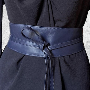 Pode incluir: Um cinto de couro azul marinho amarrado em laço, realçando um vestido escuro. O cinto tem uma textura lisa e uma gravata longa. O vestido tem mangas compridas e um decote franzido.