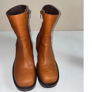 May include: A pair of mid-calf boots in a tan leather finish featuring a square toe design, a thick black lug sole, and a vertical zipper closure on the inner side of each boot. The interior lining of the boots is a dark brown color.
