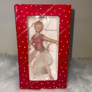 May include: A ballerina ornament in a red box with white polka dots. The ballerina wears a pink bodice, a white tutu, and pink ballet slippers. The box reads "Dillard's Trimmings".