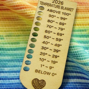 May include: A wooden temperature blanket guide with the text "2026 TEMPERATURE BLANKET" and temperature ranges from "ABOVE 100°" to "BELOW 0°". The guide has a heart-shaped yarn design at the bottom and is set against a colorful, striped background.