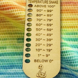 May include: A wooden temperature gauge labeled "2026 TEMPERATURE SNAKE" with temperature ranges from above 100°F to below 0°F. The gauge has a snake illustration at the bottom and is set against a colorful, rainbow-striped background.