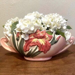May include: A pale pink ceramic bowl with two handles, featuring a raised relief design of a large red and yellow iris flower with green leaves. The bowl is filled with a cluster of white hydrangea flowers with green leaves visible among the petals.