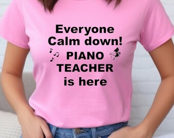 May include: A light pink t-shirt with the text "Everyone Calm down! PIANO TEACHER is here" in black. Musical notes and a piano teacher graphic are also included. The shirt is short-sleeved and made of a soft material.