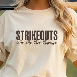 May include: A beige t-shirt with the text "STRIKEOUTS Are My Love Language" in dark brown lettering. The shirt is made of a soft material and has a crew neck. The person is wearing blue jeans.