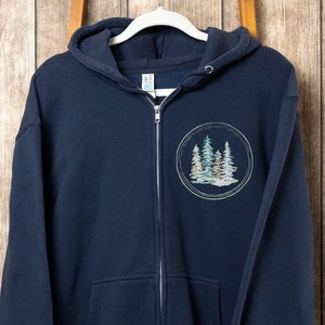 May include: Navy blue zippered hoodie with a circular design featuring a group of stylized trees in shades of green, brown, and white. The text "Color: Navy" is printed on the bottom of the hoodie.