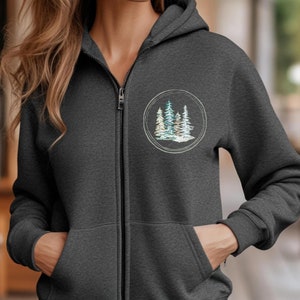 May include: A dark gray zip-up hoodie with a pocket on the front. The hoodie features a circular design with a watercolor-style image of three pine trees.