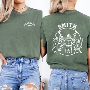 Puede incluir: Camiseta verde con el texto "Baseball Mom" en la parte delantera y un dise&ntilde;o de tem&aacute;tica de b&eacute;isbol en la parte posterior. El dise&ntilde;o presenta a tres jugadores de b&eacute;isbol con sus nombres y n&uacute;meros: Jake 1, Jacob 2 y John 3. El texto "Smith" est&aacute; por encima del dise&ntilde;o.