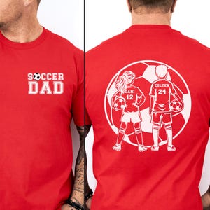 May include: Red t-shirt with white text that says "SOCCER DAD" on the front. The back of the shirt features a white graphic of two soccer players inside a soccer ball, with the names "DANI 12" and "COLTEN 24".