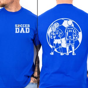 May include: Royal blue t-shirt with white text and graphics. The front displays "SOCCER DAD" in a bold font. The back features a soccer ball graphic with two children, "DANI 12" and "COLTEN 24", holding soccer balls.
