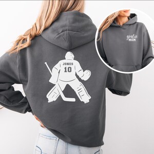 Op de afbeelding: Donkergrijze hoodie met een witte hockey goalie afbeelding op de rug, met de naam "JONES" en het nummer "10". Een kleinere afbeelding toont de tekst "goalie MOM" op de voorkant.