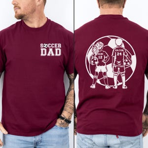 May include: Burgundy t-shirt with the text "SOCCER DAD" in white on the front. The back features a white graphic of two soccer players with the names "DANI 12" and "COLTEN 24" inside a soccer ball.