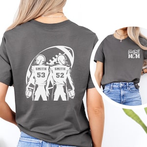 May include: Gray t-shirt featuring a football-themed graphic. The back of the shirt displays two football players with the name "Smith" and numbers 53 and 52. The front of the shirt has the text "football MOM".