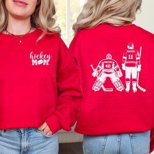 May include: Red sweatshirt with the text "hockey mom" and a hockey puck on the front. The back of the sweatshirt has a white silhouette of two hockey players, one is a goalie and the other is a forward.