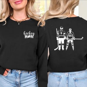 May include: Black sweatshirt with the text "hockey MOM" in white script and a hockey puck graphic. The back of the sweatshirt features a white graphic of two hockey players with the name "Smith" and numbers 5 and 55.