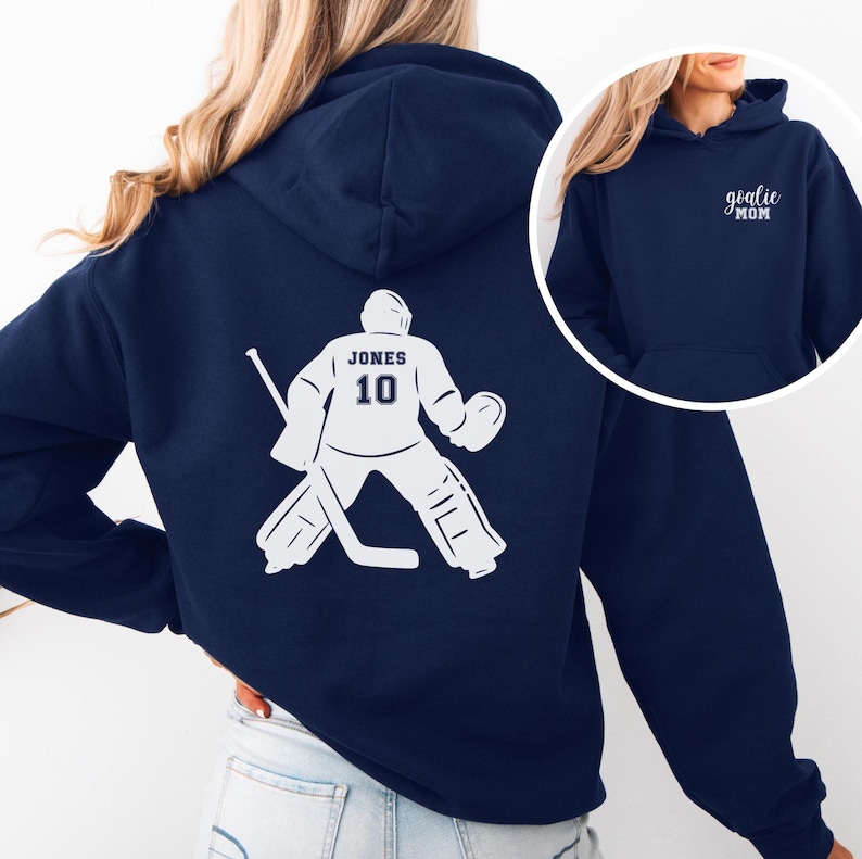 Op de afbeelding: Marineblauwe hoodie met een witte hockey goalie graphic op de rug, met de naam "JONES" en het nummer "10". De voorkant van de hoodie heeft de tekst "goalie MOM" in het wit.