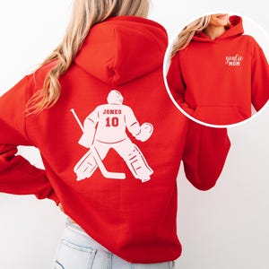Op de afbeelding: Een rode hoodie met een witte hockey goalie graphic op de rug, met de naam "JONES" en het nummer "10". De voorkant van de hoodie heeft de tekst "goalie MOM" in het wit.
