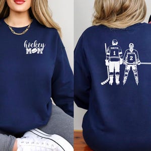 May include: Navy blue sweatshirt with white text and graphics. The front features the words "hockey mom" with a hockey puck graphic. The back displays two hockey players with the name "Smith" and numbers 1 and 2.