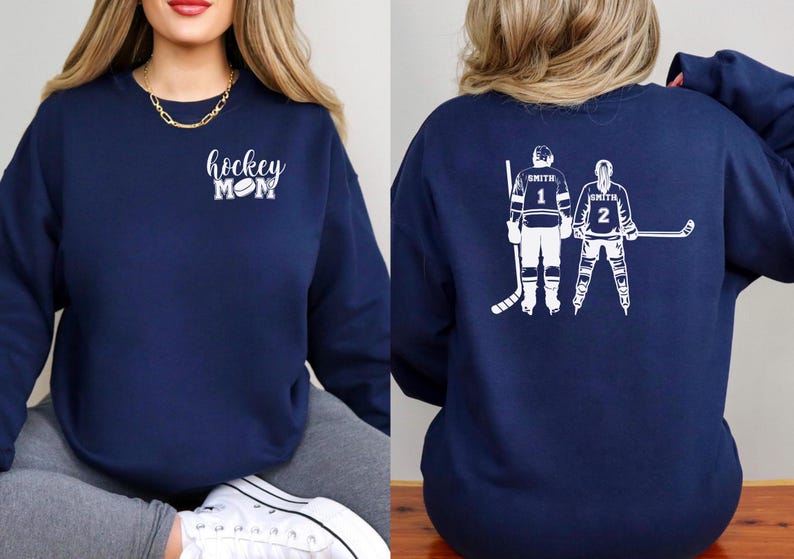May include: Navy blue sweatshirt with white text and graphics. The front features the words "hockey mom" with a hockey puck graphic. The back displays two hockey players with the name "Smith" and numbers 1 and 2.