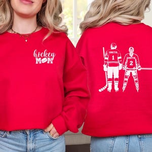May include: Red sweatshirt with white text reading "hockey MOM" and a hockey puck graphic. The back displays a white graphic of two hockey players with the name "Smith" and the numbers 1 and 2.