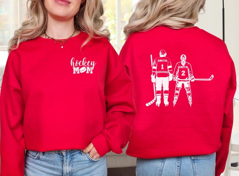 May include: Red sweatshirt with white text reading "hockey MOM" and a hockey puck graphic. The back displays a white graphic of two hockey players with the name "Smith" and the numbers 1 and 2.