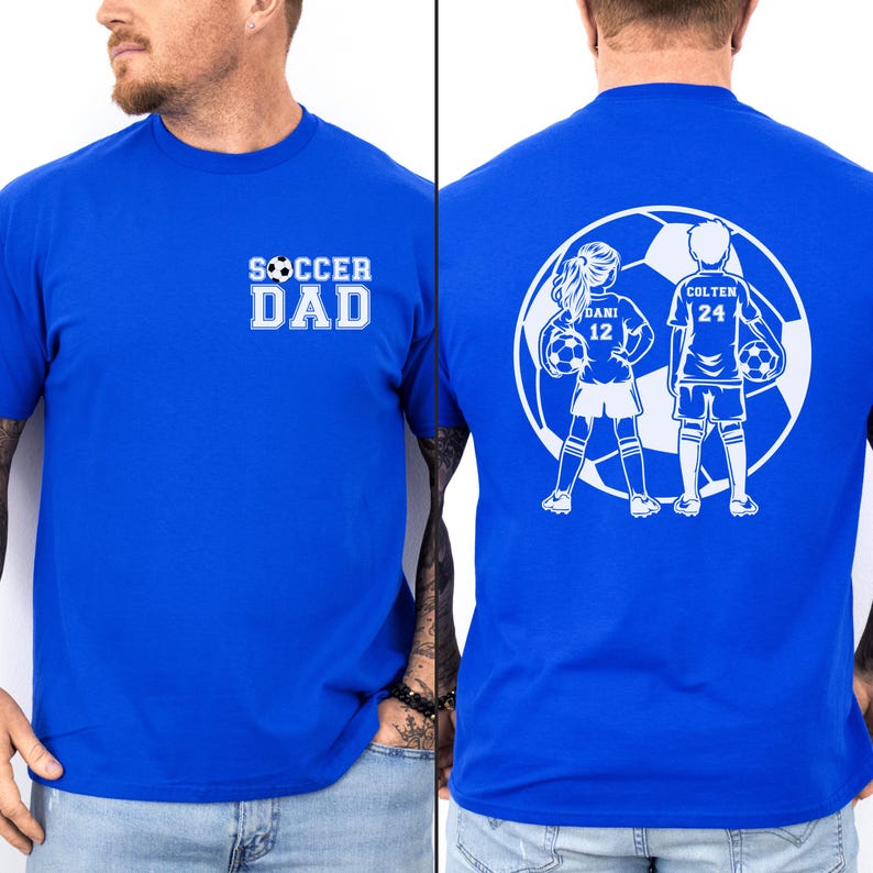 May include: Royal blue t-shirt with white text and graphics. The front displays "SOCCER DAD" in a bold font. The back features a soccer ball graphic with two children, "DANI 12" and "COLTEN 24", holding soccer balls.