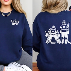 May include: Two navy blue sweatshirts. One sweatshirt has the text "hockey mom" with a hockey puck on the left chest. The other sweatshirt has a white graphic of two hockey players on the back. One player is a goalie with the number 40 and the other player has the number 11.