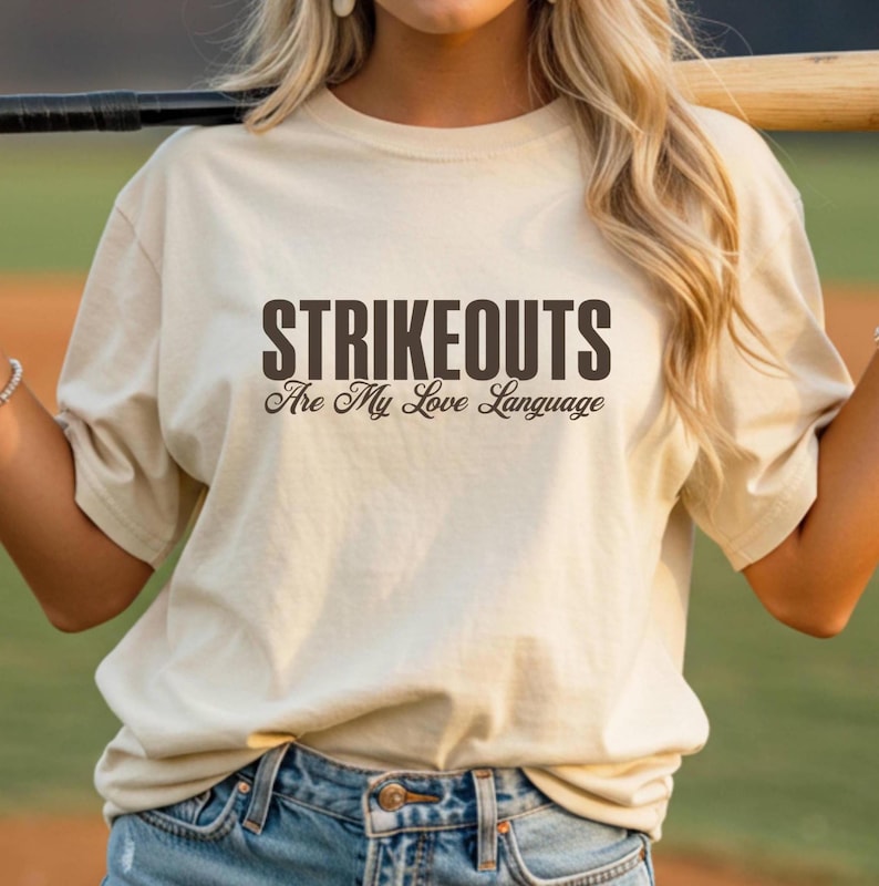 May include: A beige t-shirt with the text "STRIKEOUTS Are My Love Language" in dark brown lettering. The shirt is made of a soft material and has a crew neck. The person is wearing blue jeans.