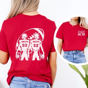 May include: Red t-shirt with a white graphic of two football players inside a football shape. The players have the name "Smith" and numbers 53 and 52. A smaller image shows a red t-shirt with "football MOM" text.